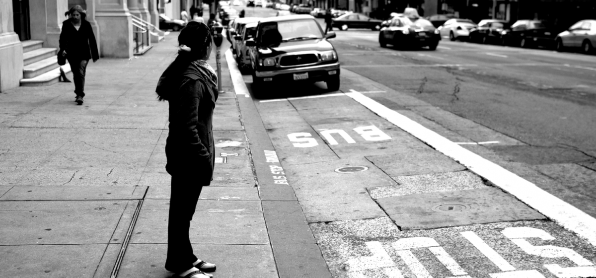 Black and white photo of person facign away from camera waiting at an empty bus stop